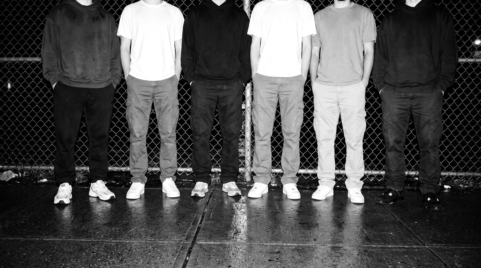 Row of six people in plain oversized hoodies and tees, photographed from the chest down on a city street at night